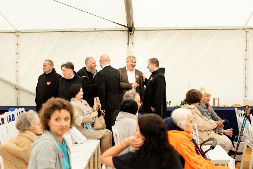 Grupa zakonników Bonifratrów oraz zaproszeni goście w eleganckich strojach rozmawiają i uśmiechają się w przestrzeni namiotowej. W tle widać osoby siedzące, w tym seniorów i kobiety, niektórzy z kijkami do chodzenia. Na twarzach wielu osób przypięte czerwone serduszka – symbol wydarzenia
