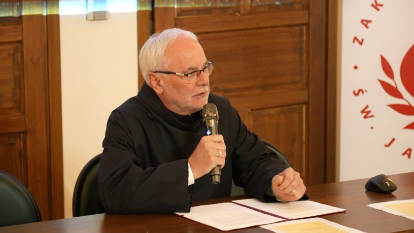 Zbliżenie na zakonnika w czarnym habicie siedzącego za stołem. Mężczyzna trzyma mikrofon i mówi, przed nim rozłożone dokumenty. W tle drewniane drzwi i fragment banera z logo Zakonu Szpitalnego św. Jana Bożego