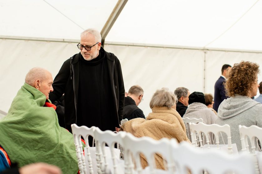 Zakonnik w czarnym habicie pochyla się w kierunku starszego mężczyzny siedzącego na wózku inwalidzkim, okrytego zielonym i czerwonym kocem. Wokół nich siedzą inni seniorzy, również otuleni kocami, a całe spotkanie odbywa się pod namiotem