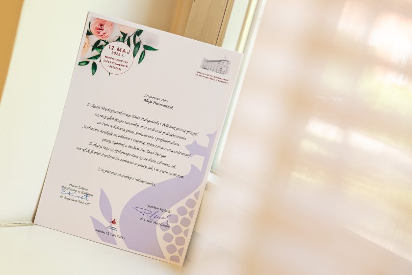 Estetycznie sfotografowana karta gratulacyjna oparta o okno, z widocznym nadrukiem „12 MAJ 2025 r.” i personalizowaną treścią skierowaną do pani Alicji Ćwiorawczyk. Po obu stronach listu – podpisy Przeora Zakonu oraz Dyrektora Szpitala. W tle miękkie, rozproszone światło zza firany, budujące spokojny, pełen szacunku nastrój