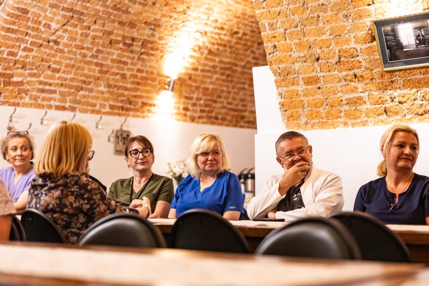 Widok drugiej części sali – kilka kobiet w różnokolorowych uniformach oraz lekarz w białym kitlu. Atmosfera skupienia, rozmów i oczekiwania na dalszą część uroczystości. Na tle ceglanych murów i jasnych ścian postacie zyskują szczególną wyrazistość.