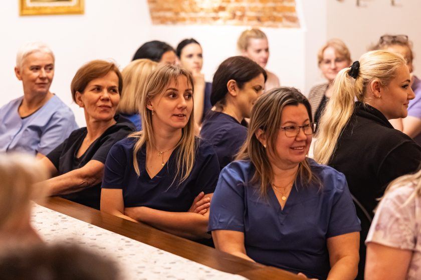 Grupa kobiet, głównie w medycznych uniformach w odcieniach niebieskiego i fioletu, uważnie słucha wystąpienia. Zdjęcie wykonane w sali z ceglanym sklepieniem i obrazem religijnym na ścianie, co podkreśla historyczny i duchowy wymiar przestrzeni. Wyraz skupienia i refleksji na twarzach uczestniczek.