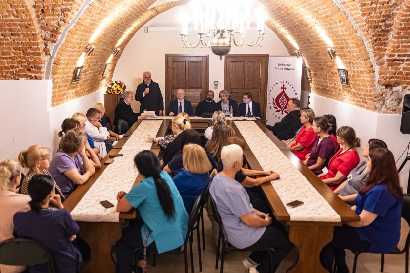 Panorama całej sali – długa sala z łukowatym ceglanym sklepieniem, uczestnicy zasiadający po obu stronach stołu. W centrum przemawia zakonnik z mikrofonem. Na stole w tle stos kartek gratulacyjnych. Uroczysty nastrój, podniosłość chwili i szacunek wobec zebranych pielęgniarek