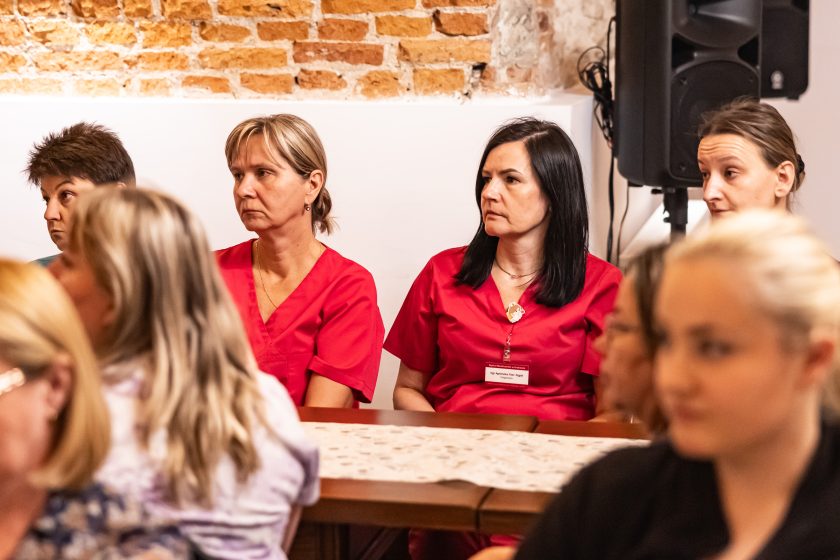 Fragment grupy kobiet w czerwonych uniformach siedzących pod ścianą. W centrum uwagę zwraca pielęgniarka z identyfikatorem – wyraz zadumy i powagi na twarzach uczestniczek. Ujęcie oddaje skupienie oraz atmosferę refleksji nad trudną, ale szlachetną profesją