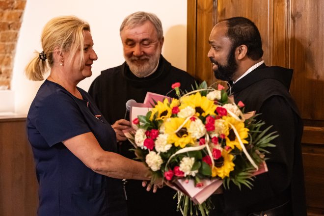 Wzruszający moment wręczenia bukietu kwiatów – uśmiechnięta pielęgniarka w granatowym uniformie przyjmuje okazały bukiet z rąk brata zakonnego o ciemnej karnacji, ubranego w czarny habit. W tle starszy zakonnik z siwą brodą, trzymający mikrofon i dyplomy, z serdecznym uśmiechem obserwuje scenę. Ciepła atmosfera, gest wdzięczności i uznania dla pracy personelu medycznego