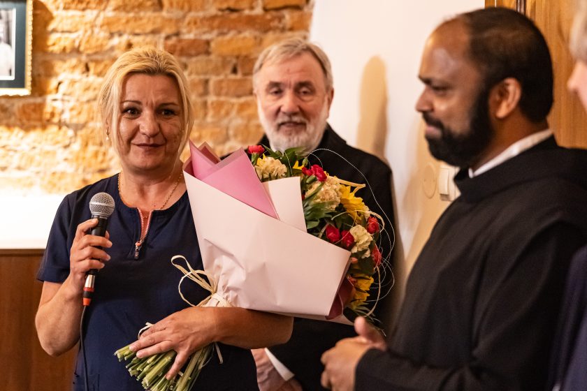 Ujęcie przedstawia uśmiechniętą pielęgniarkę w granatowym uniformie trzymającą mikrofon oraz okazały bukiet kwiatów z żółtymi słonecznikami i czerwonymi różami, starannie opakowanymi w papier z różowym akcentem. Kobieta zwraca się do zebranych, najprawdopodobniej dziękując za wyróżnienie. Za nią stoi zakonnik z siwą brodą, a po prawej stronie kadr domyka brat zakonny w czarnym habicie. Ciepły klimat pomieszczenia i ceglana ściana tworzą przyjazne tło dla wzruszającej chwili