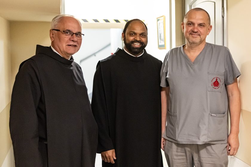 Br. Saji Mullankuzhy stoi pomiędzy dwoma mężczyznami – jednym w habicie, drugim w szarym uniformie medycznym z logo szpitala. Trójka pozuje do wspólnego zdjęcia w korytarzu placówki. Ujęcie emanuje serdecznością i współpracą między zakonem a personelem medycznym