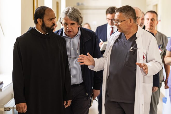 Trójka mężczyzn – br. Saji, lekarz i przedstawiciel dyrekcji – prowadzi intensywną rozmowę podczas przemierzania szpitalnego korytarza. Lekarz w białym fartuchu gestykuluje, jakby tłumaczył ważną kwestię, co wskazuje na charakter roboczy wizyty