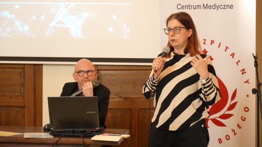 Kobieta w paskowanej bluzie i okularach przemawia do mikrofonu, stojąc na tle banera Bonifraterskiego Centrum Medycznego. Obok niej siedzi mężczyzna w okularach, ubrany na czarno, z laptopem przed sobą. W tle ekran z grafiką sieci neuronowej
