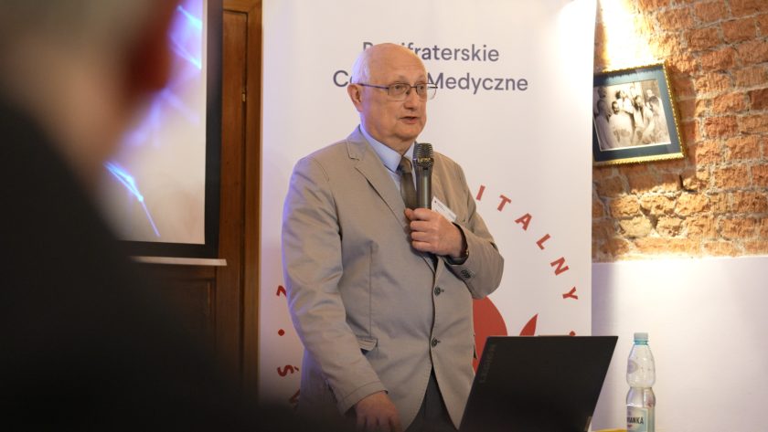 Mężczyzna w jasnym garniturze przemawia do mikrofonu, stojąc przy laptopie na tle banera Bonifraterskiego Centrum Medycznego. W tle ceglana ściana z oprawioną fotografią. Na stole butelka wody i myszka komputerowa