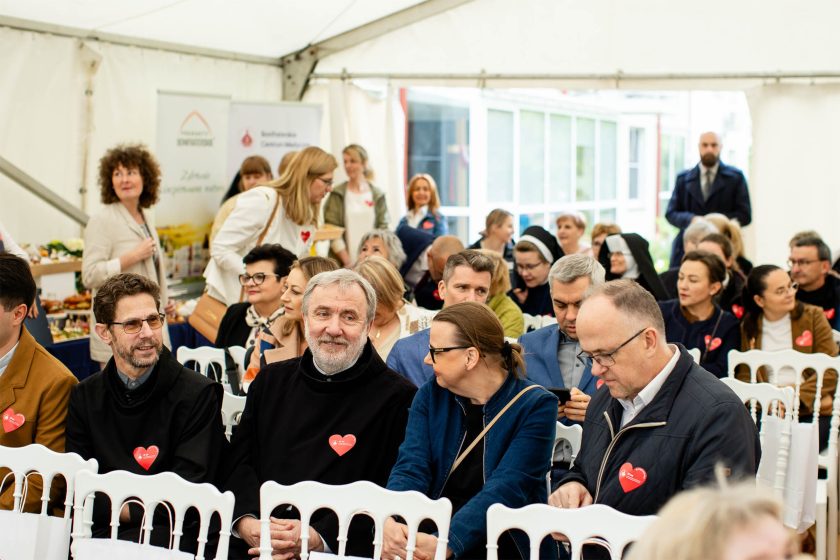 Widok na tył namiotu z uczestnikami wydarzenia. W pierwszym rzędzie zakonnik w czarnym habicie uśmiecha się do osób obok, w tle widać siostry zakonne i personel, a po lewej stronie banery informacyjne z logotypami Bonifraterskiego Centrum Medycznego i Produktów Bonifraterskich