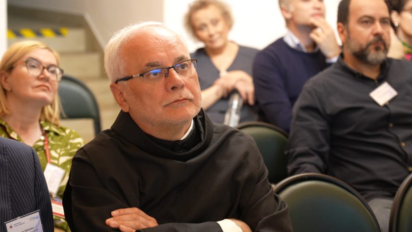 Zbliżenie na zakonnika w habicie siedzącego wśród uczestników konferencji. Mężczyzna patrzy uważnie na prelegenta. W tle inne osoby, w tym kobieta z butelką wody i mężczyzna w granatowym swetrze