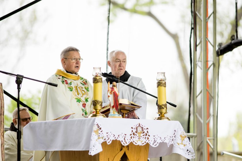 Dwóch księży odprawia mszę świętą na scenie – jeden w białej szacie z kolorowym haftem, drugi w albie. Przed nimi ołtarz z figurką Zmartwychwstałego Jezusa, świecami i obrusem z haftowanym motywem religijnym. W tle zieleń i fragment konstrukcji scenicznej