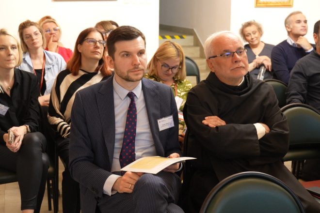 Ujęcie szerokie na grupę uczestników siedzących w sali. W pierwszym rzędzie zakonnik i mężczyzna w garniturze z kartką w ręku. Obok kobieta w paskowanej bluzie. W tle kolejne rzędy słuchaczy śledzących przebieg spotkania
