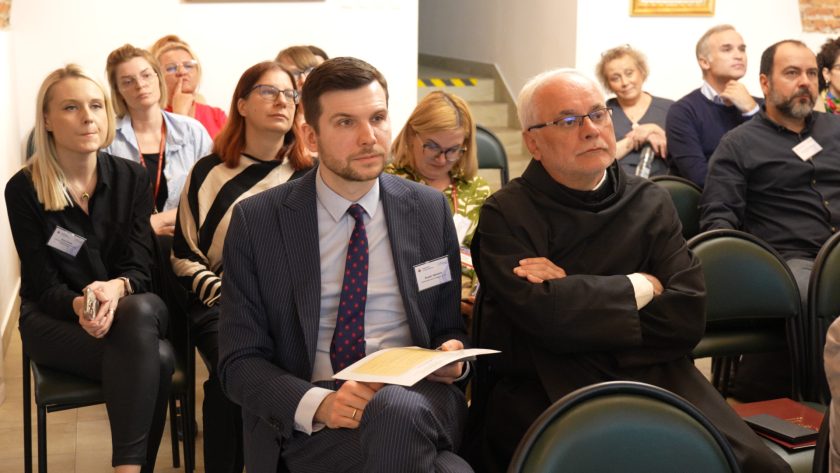 Ujęcie szerokie na grupę uczestników siedzących w sali. W pierwszym rzędzie zakonnik i mężczyzna w garniturze z kartką w ręku. Obok kobieta w paskowanej bluzie. W tle kolejne rzędy słuchaczy śledzących przebieg spotkania