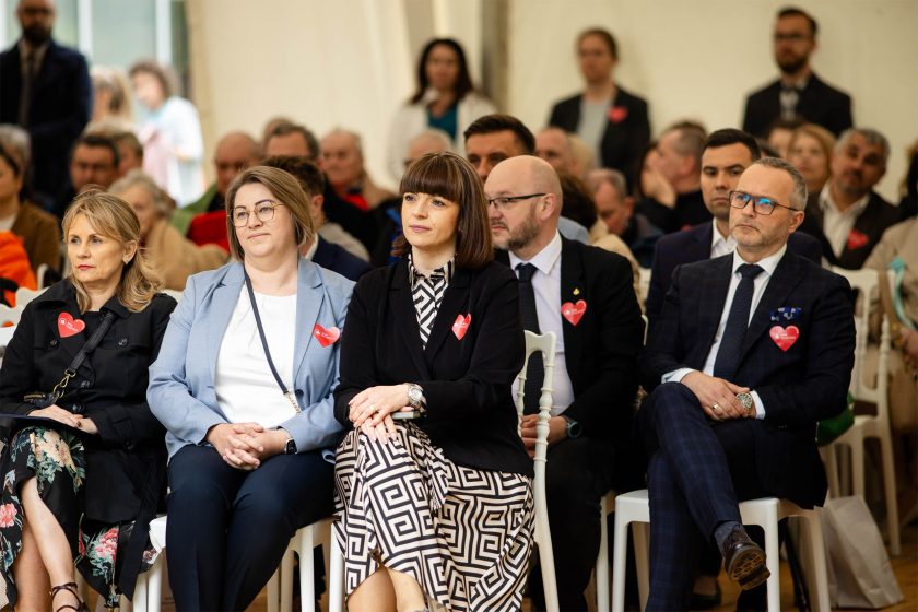 Pierwszy rząd gości siedzących w namiocie – widoczne skupienie na twarzach kobiet i mężczyzn w eleganckich strojach. Wszyscy mają przypięte czerwone serduszka. W tle tłum zgromadzonych gości – osoby świeckie i duchowne