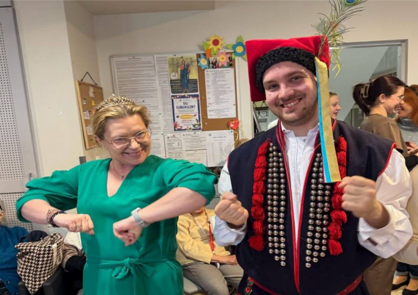 Dwie osoby pozują wesoło do zdjęcia – kobieta w zielonej sukience z koroną na głowie robi zabawny gest łokciami, a obok niej mężczyzna w stroju ludowym z czerwonym beretem z piórkiem i ozdobnym kaftanem. Za nimi osoby starsze siedzą na krzesłach, a ściana ozdobiona jest informacjami i plakatami, w tym afiszem balu karnawałowego