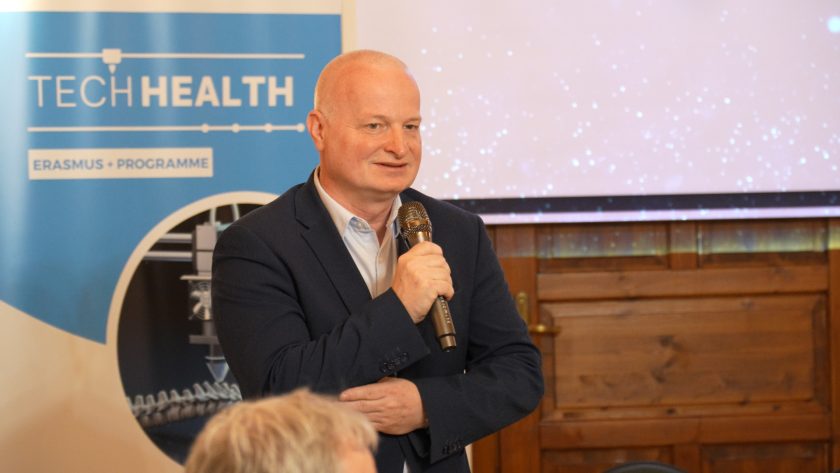 Zbliżenie na mężczyznę z mikrofonem w ręku, ubranego w granatowy garnitur i błękitną koszulę. Przemawia stojąc obok banera projektu TECHHEALTH. W tle widoczna część ekranu multimedialnego i drewniane drzwi