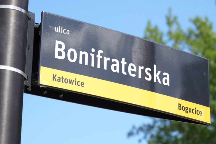 Zbliżenie na nową tablicę z nazwą ulicy – „Bonifraterska” – w Katowicach, w dzielnicy Bogucice. Żółto-czarno-biała tablica osadzona na czarnym słupie kontrastuje z błękitem nieba i zielenią liści w tle. Nazwa ulicy jest formą uhonorowania działalności Zakonu Bonifratrów w regionie, który od 150 lat świadczy pomoc medyczną i społeczną mieszkańcom miasta