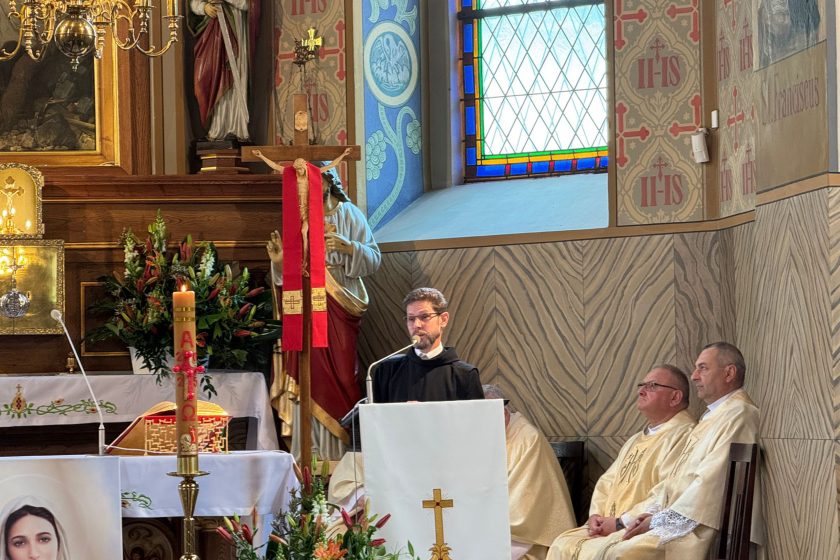 Zakonnik w czarnym habicie wygłasza homilię z ambony wewnątrz zdobionego wnętrza kościoła. Za nim, przy ścianie i ołtarzu, siedzi dwóch księży w kremowych ornatach. Otoczenie wypełniają witraże, malowane ściany z motywami liturgicznymi i kwiaty ustawione przy ołtarzu. Przed amboną znajduje się świeca paschalna z napisem „AΩ 2024”. Atmosfera zdjęcia jest podniosła i skupiona, a całe wnętrze podkreśla uroczysty charakter wydarzenia