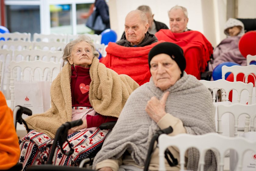 Grupa starszych osób siedzących na wózkach inwalidzkich, otulonych kocami w czerwonym, beżowym i szarym kolorze. Uczestniczą w uroczystości pod namiotem, uważnie obserwując wydarzenie. W tle biało-czerwone balony i rzędy pustych białych krzeseł