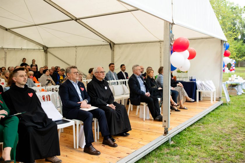 Szeroki kadr na wnętrze namiotu z uczestnikami siedzącymi na białych krzesłach. Na pierwszym planie osoby w habitach i garniturach, z boku widoczne czerwono-biało-niebieskie balony i stół z materiałami. Po bokach widać zieleń ogrodu