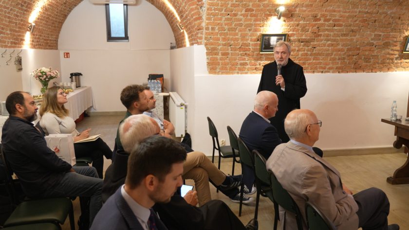 Szerokie ujęcie sali konferencyjnej z widokiem na grupę uczestników siedzących frontem do zakonnika przemawiającego z mikrofonem. W tle ceglane sklepienie i stolik z poczęstunkiem. Atmosfera skupienia i zaangażowania