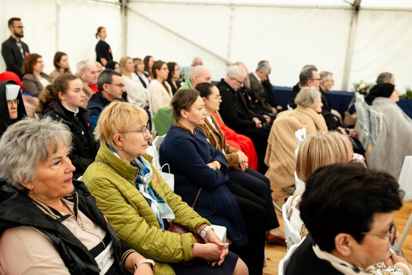 Publiczność siedząca w skupieniu podczas uroczystości. Na pierwszym planie kobiety w kurtkach i szalikach, w tle m.in. osoby duchowne, wolontariusze i goście
