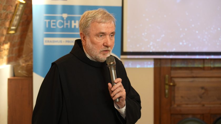 Zbliżenie na zakonnika z siwą brodą, przemawiającego do mikrofonu na tle banera TECHHEALTH. Mężczyzna w czarnym habicie spogląda w stronę uczestników z powagą