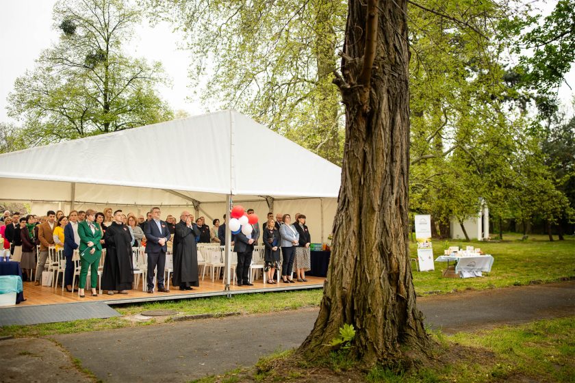 Ogólny widok na namiot z uczestnikami wydarzenia stojącymi wewnątrz. Na pierwszym planie widoczny pień drzewa, w tle zielony teren z punktami informacyjnymi i stoiskami promocyjnymi