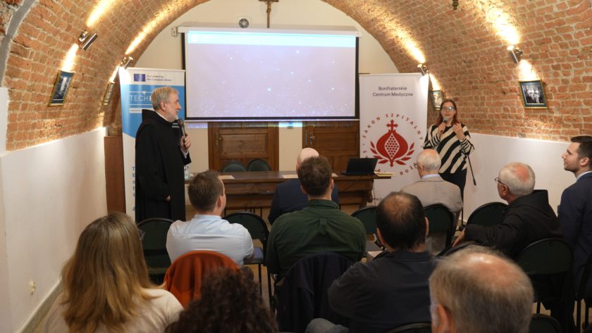 Sala konferencyjna o sklepionym ceglanym suficie. Przy stole prezydialnym siedzi zakonnik w czarnym habicie, przemawiający do mikrofonu. Obok stoi kobieta w okularach i biało-czarnej bluzie w paski, również trzymająca mikrofon. Za nimi rozwieszone są dwa roll-upy: jeden z logo projektu TECHHEALTH i drugi z logo Bonifraterskiego Centrum Medycznego