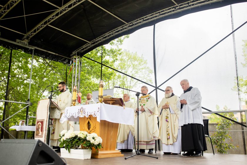 Ołtarz polowy na scenie, przy którym zgromadzonych jest kilku księży i zakonników w liturgicznych szatach. W centrum kadru ksiądz wygłasza słowa podczas mszy świętej