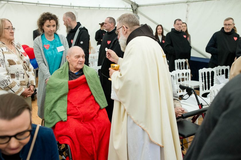 Ksiądz rozdaje komunię starszemu mężczyźnie siedzącemu na wózku, otulonemu czerwonym i zielonym kocem. W tle inni zakonnicy oraz osoby świeckie