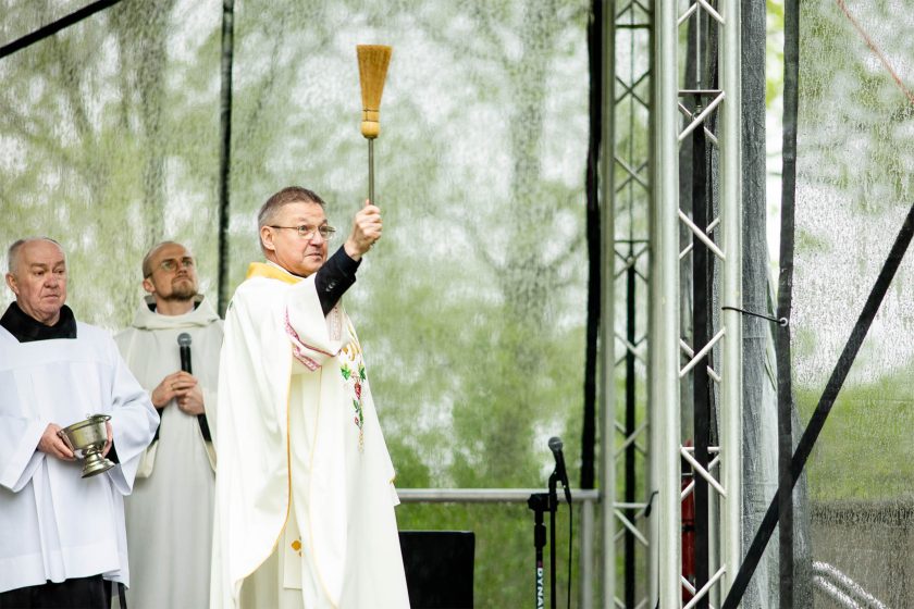 Kapłan w liturgicznej szacie błogosławi zgromadzonych, wykonując gest kropienia wodą święconą. Obok ministranci i osoby duchowne z naczyniem na wodę