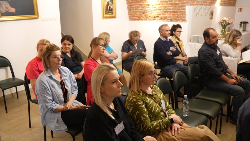 Szerszy widok grupy uczestników spotkania. W kilku rzędach siedzą kobiety i mężczyźni – pracownicy i słuchacze konferencji. W tle dekoracje z kwiatami, ceglane ściany i obrazy nawiązujące do religii i historii Zakonu