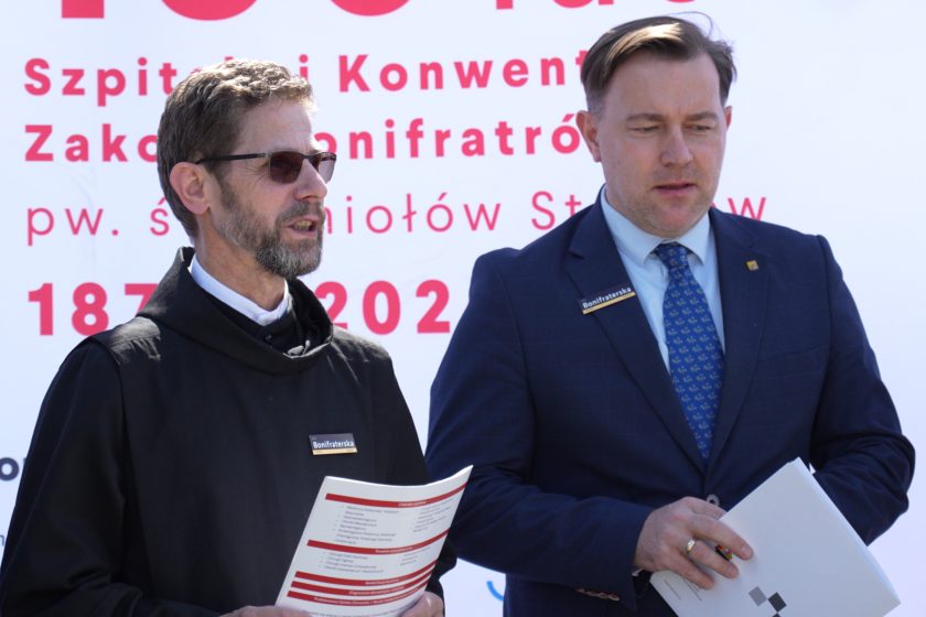 Zbliżenie na dwóch kluczowych uczestników uroczystości jubileuszowych – zakonnika w czarnym habicie z naszywką „Bonifraterska” oraz mężczyznę w garniturze, również z identyfikacją z nazwą ulicy. Obaj trzymają dokumenty związane z obchodami 150-lecia Szpitala i Konwentu Bonifratrów. W tle biało-czerwony baner z datami „1874–2024” akcentuje wagę rocznicy. Obraz oddaje powagę i znaczenie chwili, jednocześnie eksponując współczesne, profesjonalne podejście do organizacji wydarzeń o charakterze historycznym