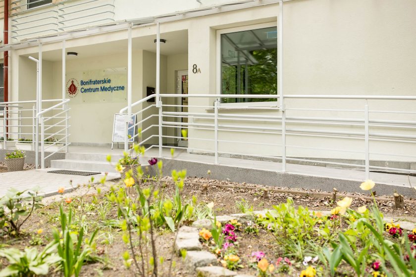 Wejście do wyremontowanego budynku Hospicjum Św. Jana Bożego przy ul. Poświęckiej 8A we Wrocławiu. Widoczny logotyp Bonifraterskiego Centrum Medycznego, podjazd z poręczami dla osób z niepełnosprawnościami oraz kwietnik z wiosennymi kwiatami w pierwszym planie