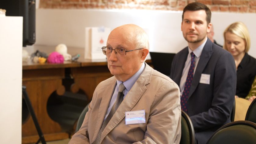 Profesor Krzysztof Gromada oraz pozostali uczestnicy w eleganckich garniturach siedzący w pierwszych rzędach sali wykładowej, z uwagą przysłuchują się prezentacji. W tle widać drewniane elementy wystroju oraz dekoracje związane z projektem