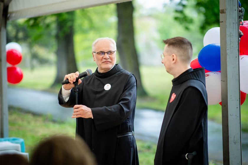 Brat bonifrater w okularach przemawia do zgromadzonych przez mikrofon, trzymając statyw, obok stoi inny brat uśmiechający się z uwagą. W tle widać zielone drzewa oraz biało-czerwono-niebieskie balony