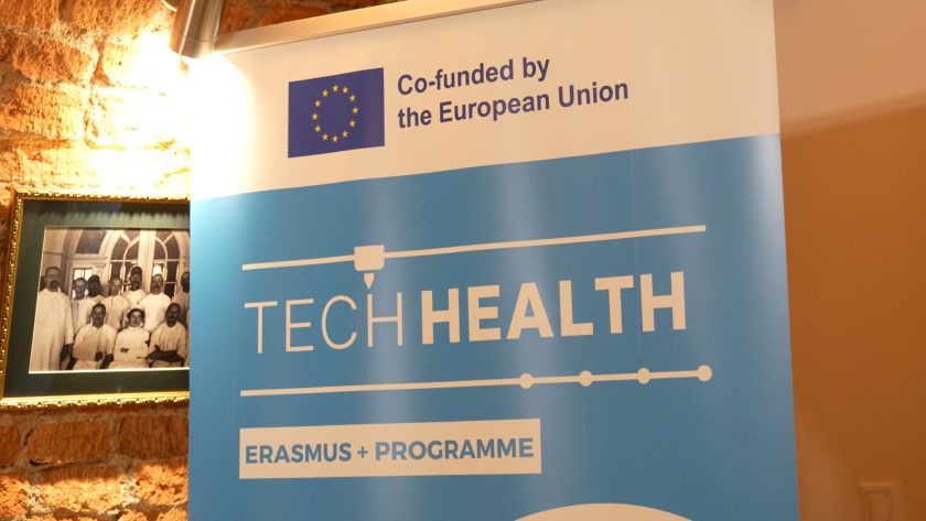 Zbliżenie na niebieski baner projektu TECHHEALTH z logo Unii Europejskiej i programu Erasmus+, ustawiony przy ceglanym murze, obok historycznej czarno-białej fotografii pracowników szpitala