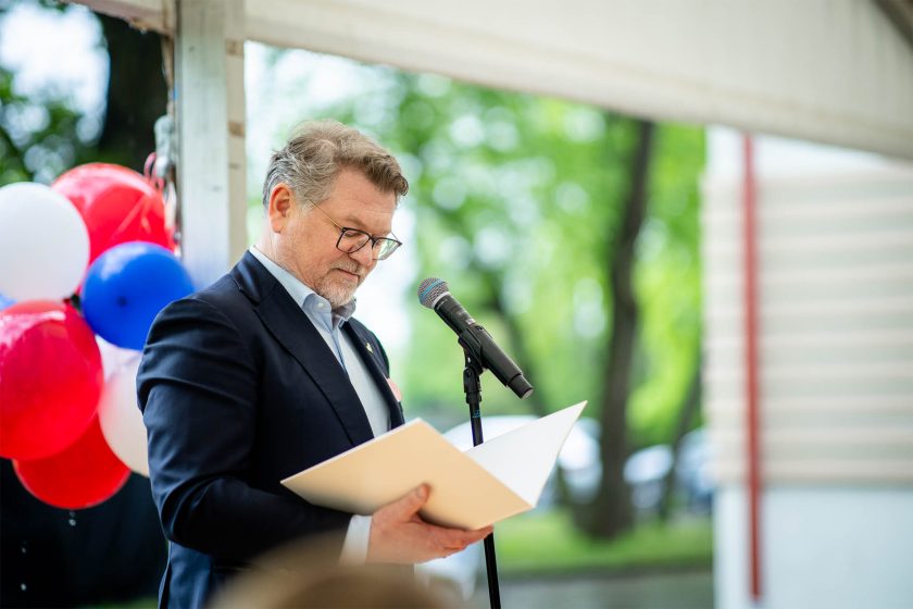 Mężczyzna w granatowej marynarce i okularach odczytuje przemówienie – Ujęcie przedstawia mężczyznę w średnim wieku przemawiającego do mikrofonu w białym namiocie, trzymającego otwartą teczkę. W tle widać czerwono-biało-niebieskie balony i zielony park