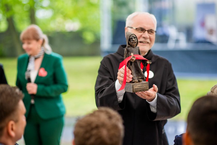 Brat zakonny prezentuje statuetkę – Mężczyzna w czarnym habicie z uśmiechem trzyma rzeźbioną figurkę zakonnika owiniętą czerwoną wstążką. W tle widoczna kobieta w zielonym garniturze i zielona przestrzeń plenerowa