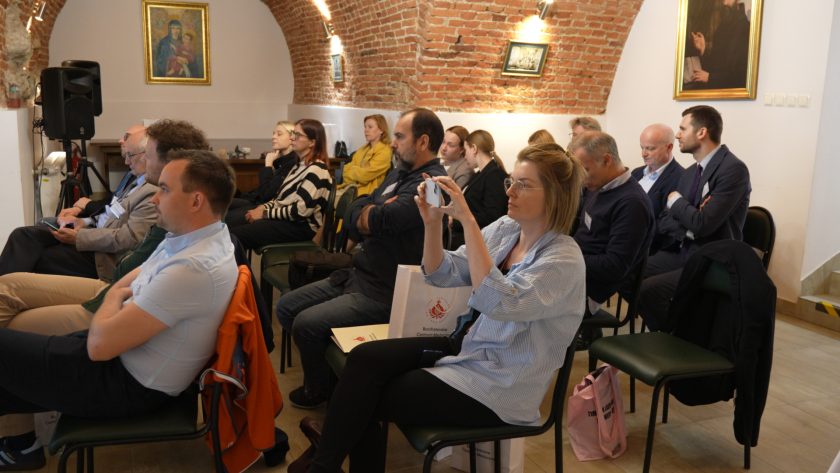 Widok ogólny na salę konferencyjną z uczestnikami słuchającymi wykładu. W pierwszym rzędzie kobieta w okularach fotografuje wydarzenie telefonem, a pozostali uczestnicy śledzą przebieg prezentacji z dużym zainteresowaniem