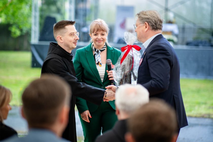 Uścisk dłoni podczas wręczania nagrody – Brat zakonny ściska dłoń mężczyzny w granatowej marynarce, który otrzymuje upominek zapakowany w celofan. Pomiędzy nimi stoi kobieta w zielonym garniturze z mikrofonem