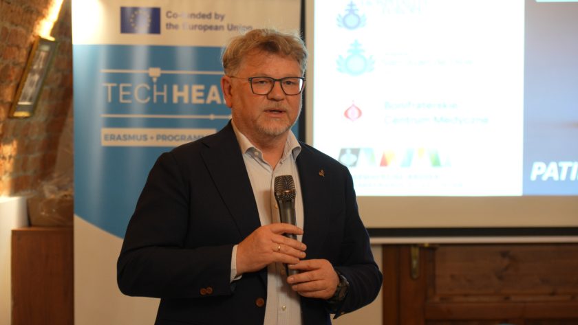 Prelegent w granatowym garniturze prezentuje temat w trakcie wystąpienia. W tle baner TECHHEALTH, ekran projekcyjny z logotypami instytucji oraz historyczny klimat ceglanych ścian sali wykładowej