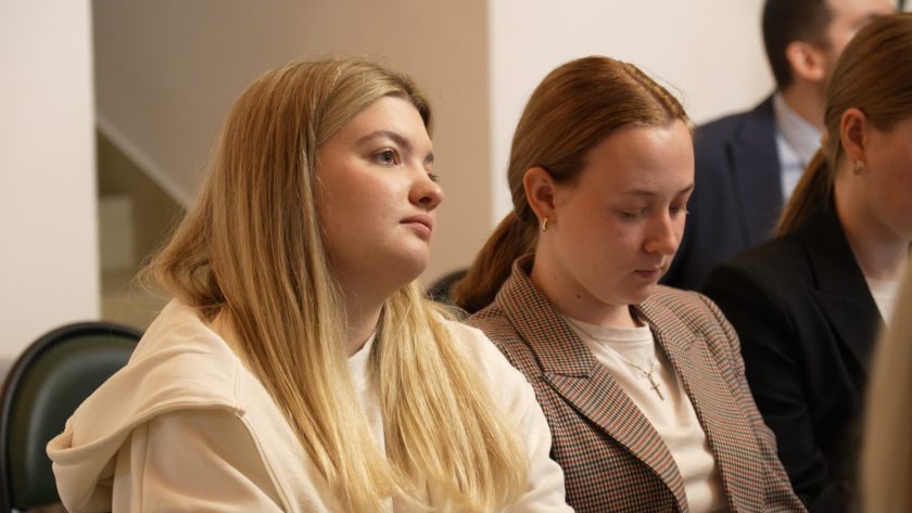 Dwie młode kobiety, jedna w jasnej bluzie, druga w kratkowanej marynarce, skupione na wykładzie. Ich spojrzenia skierowane są w stronę prelegenta, co podkreśla atmosferę zaangażowania i koncentracji