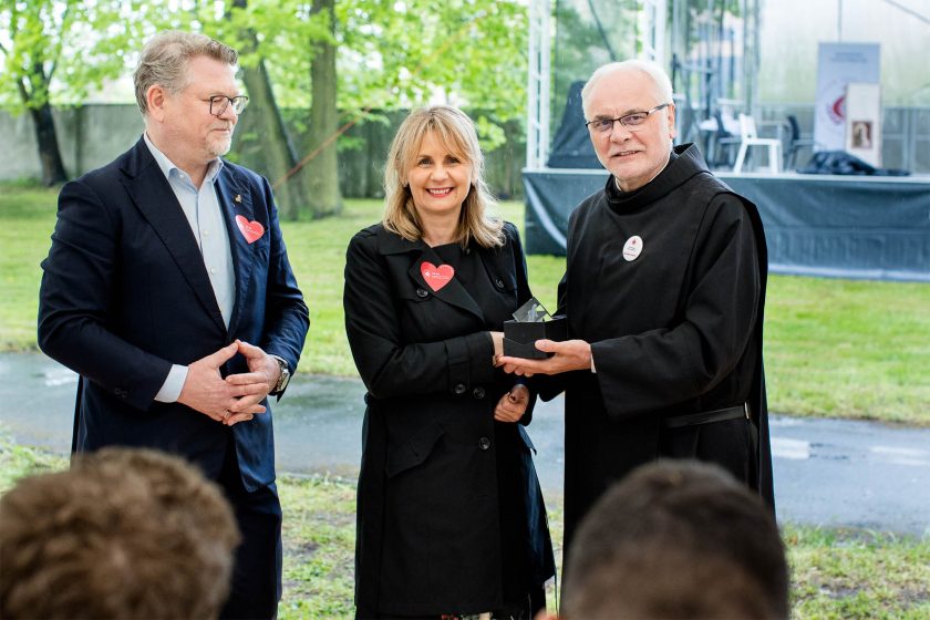 Wręczenie nagrody kobiecie w czarnym płaszczu – Brat zakonny przekazuje nagrodę uśmiechniętej kobiecie w czarnym płaszczu, stojącej obok mężczyzny w marynarce. W tle widoczny zielony park i namiot