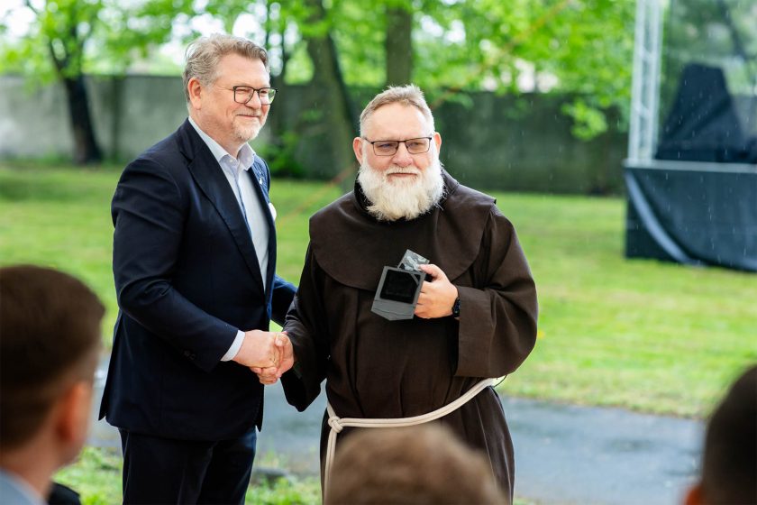 Brat zakonny i franciszkanin z nagrodą – Mężczyźni ściskają sobie dłonie, a zakonnik w brązowym habicie i z siwą brodą trzyma czarną statuetkę. Zdjęcie wykonane pod namiotem na tle deszczowego parku