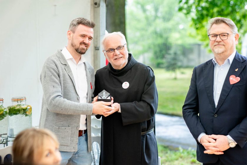 Wręczenie nagrody mężczyźnie w szarym swetrze – Brat zakonny i mężczyzna w casualowej stylizacji wspólnie trzymają statuetkę, pozując do zdjęcia. Obok nich stoi mężczyzna w marynarce z serduszkiem na klapie