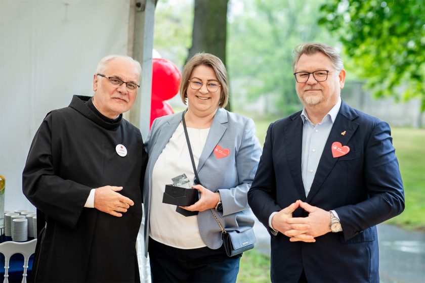 Kobieta w niebieskiej marynarce z nagrodą – Trzyosobowe zdjęcie przedstawia zakonnika, kobietę z okularami i krótkimi włosami trzymającą statuetkę oraz mężczyznę w granatowym garniturze. W tle park i namiot